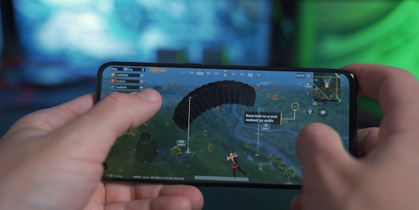 A person is holding a smartphone horizontally, playing a video game that involves a character parachuting into an open area. The screen shows various game interface elements including a compass, a mini-map, and player stats. The background has a blurred and colorful setting, suggesting a focus on the gaming experience.
